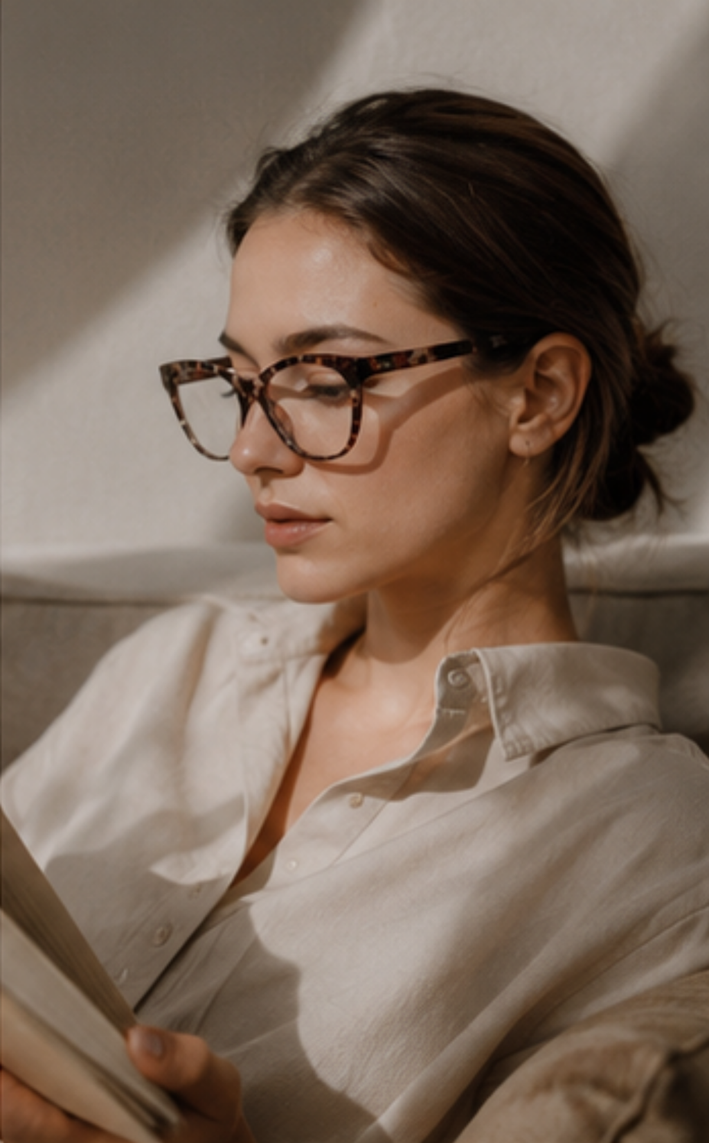 A woman with dark hair tied back wears tortoiseshell eyeglasses and a light beige button-up shirt, reading a book in a warmly lit indoor setting.