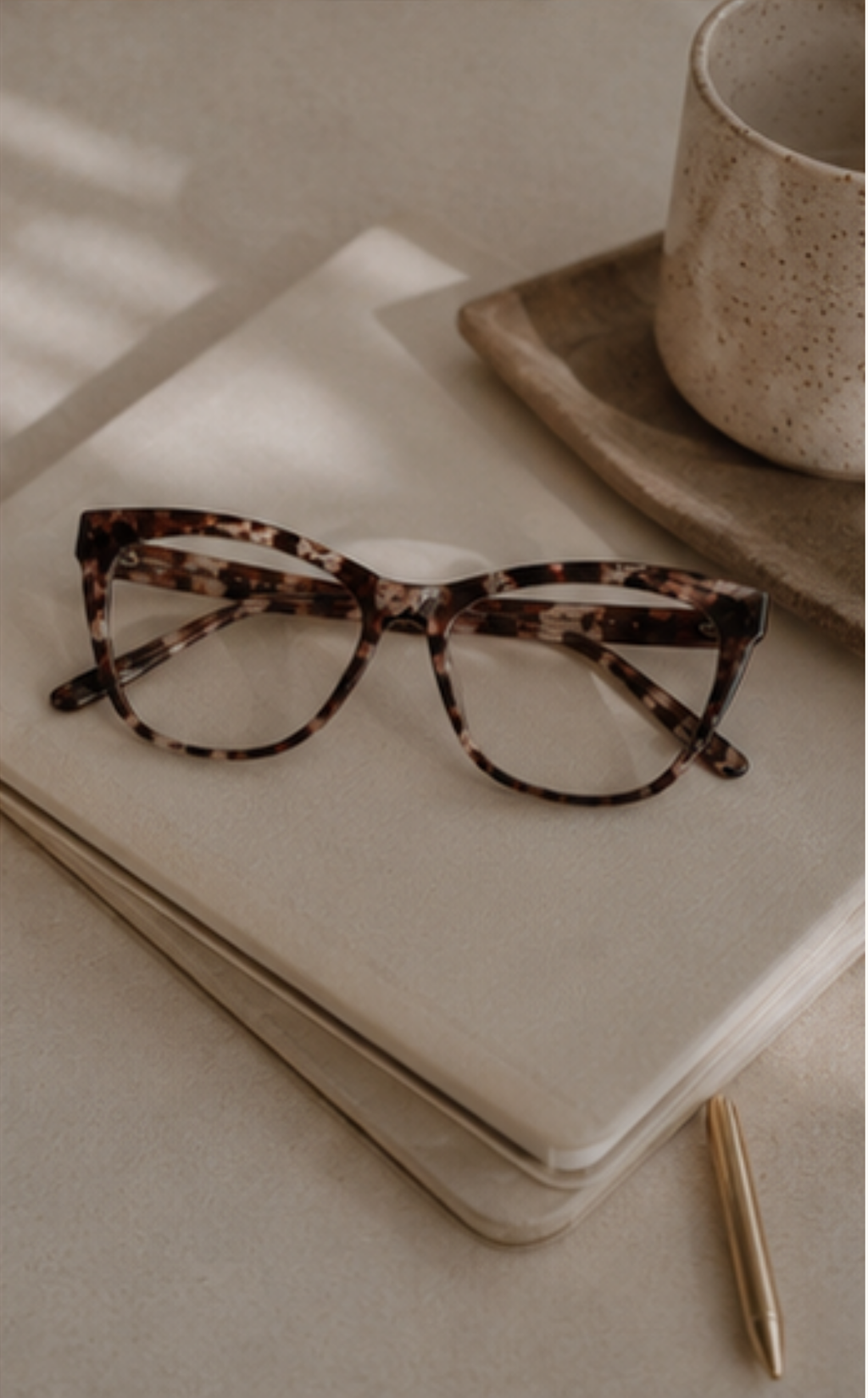 Tortoiseshell eyeglasses resting on a cream-colored notebook, with a ceramic mug on a tray and a gold pen nearby in a softly lit setting.