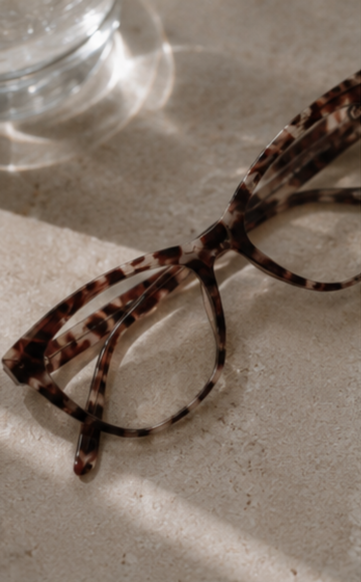 Tortoiseshell acetate eyeglasses resting on a textured beige surface with soft natural shadows.