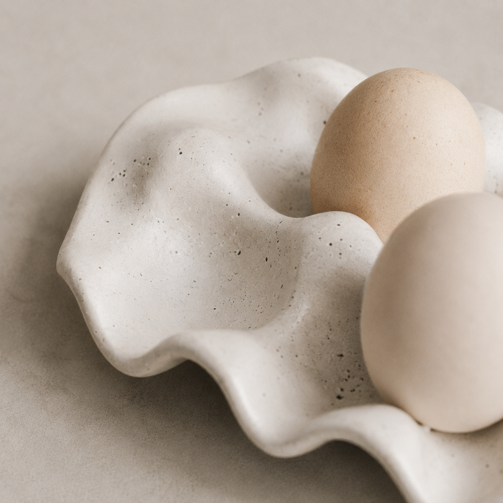 A close-up of a sculptural ceramic egg carton with two speckled eggs resting inside on a neutral beige surface.