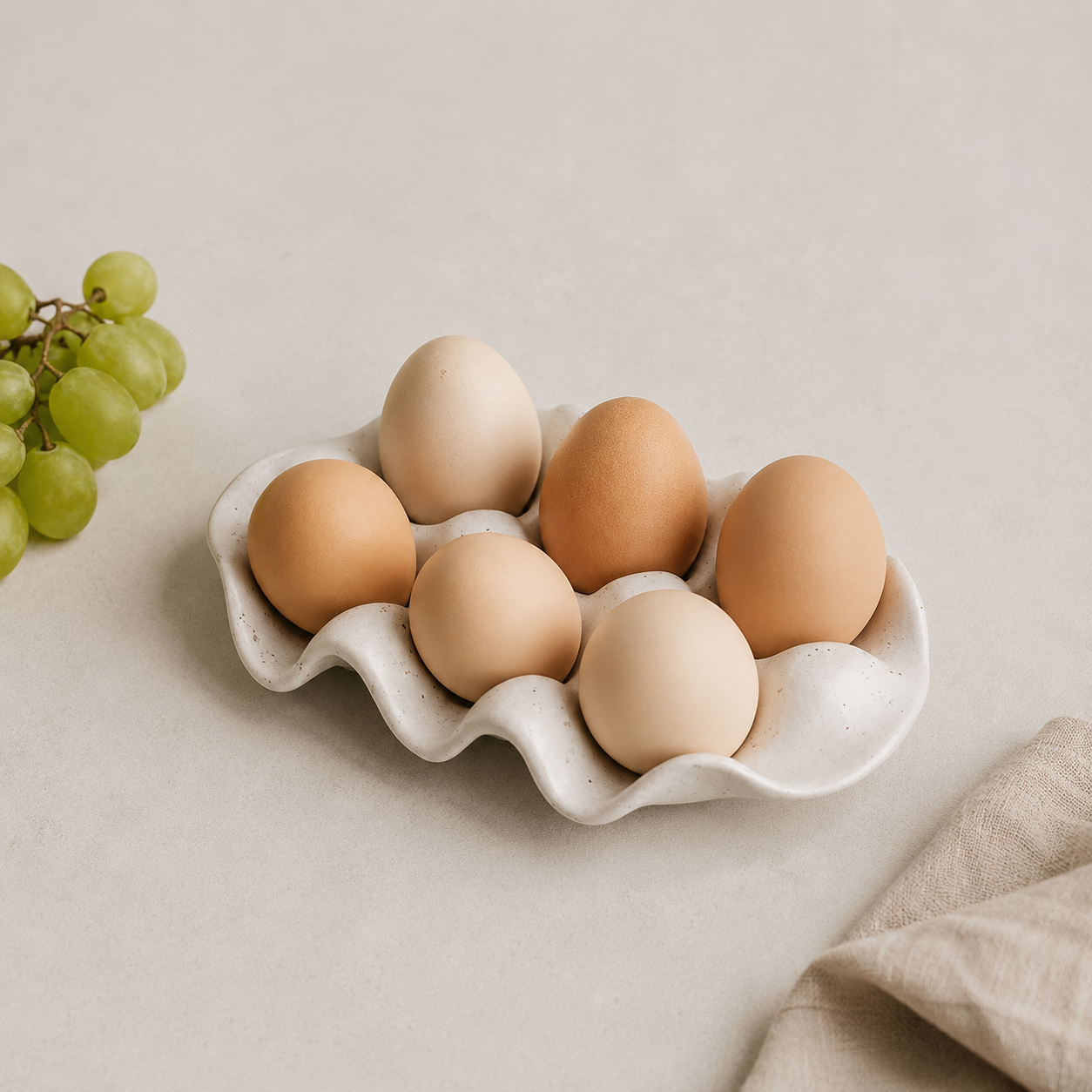 A ceramic egg tray holding six eggs, styled on a light beige surface with green grapes on the left and a beige cloth on the right.