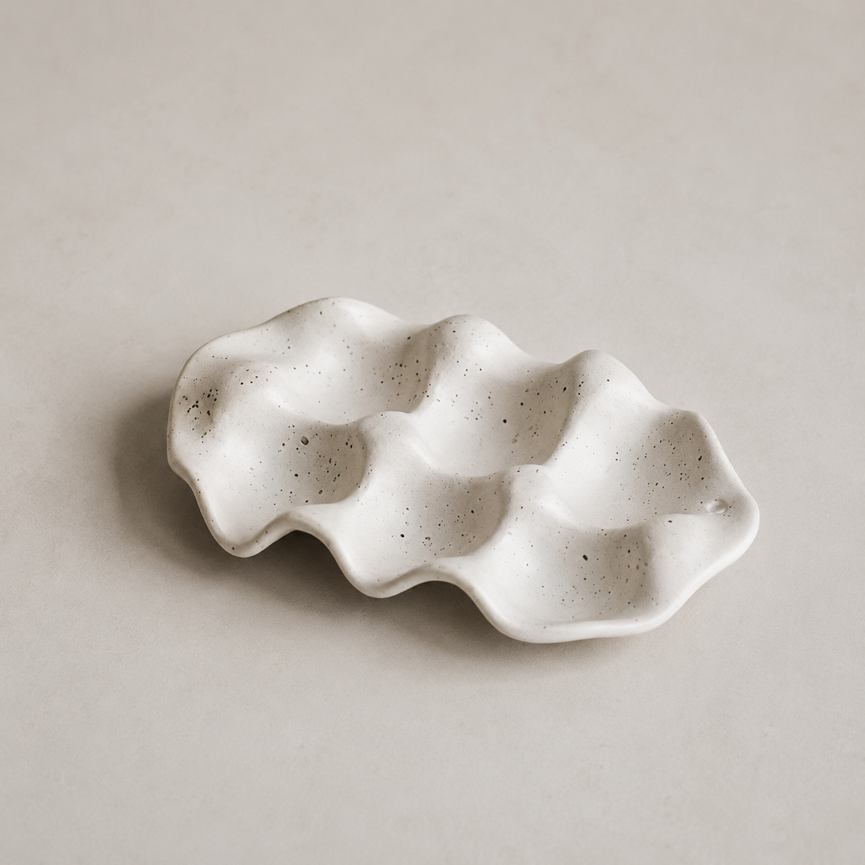 A small wavy ceramic dish with a speckled off-white finish, photographed on a neutral light beige background.