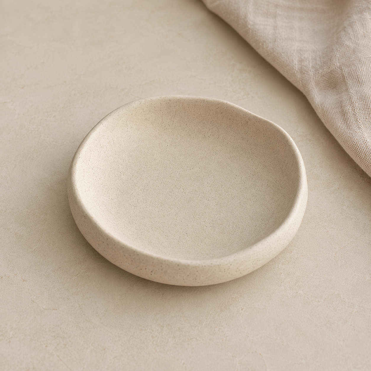 A shallow, round ceramic dish with a textured, matte finish in a warm cream-beige color, styled on a matching beige surface with a folded linen cloth in the upper right corner.