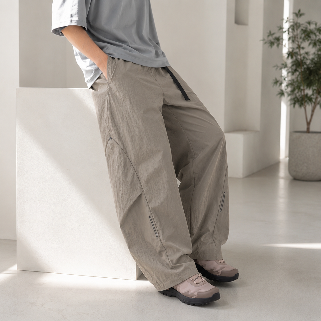 A person wearing loose, wide-leg taupe pants with a black drawstring, paired with a light gray t-shirt and brown shoes, leaning against a white block in a bright, modern minimalist room.