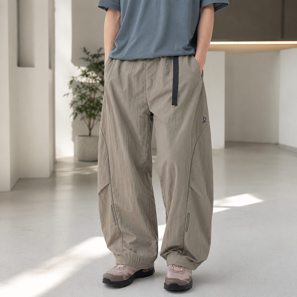A person wearing loose-fit beige cargo-style pants with a dark vertical zipper detail and elastic cuffs, paired with a muted blue-gray t-shirt and brown shoes, standing in a bright modern indoor space.