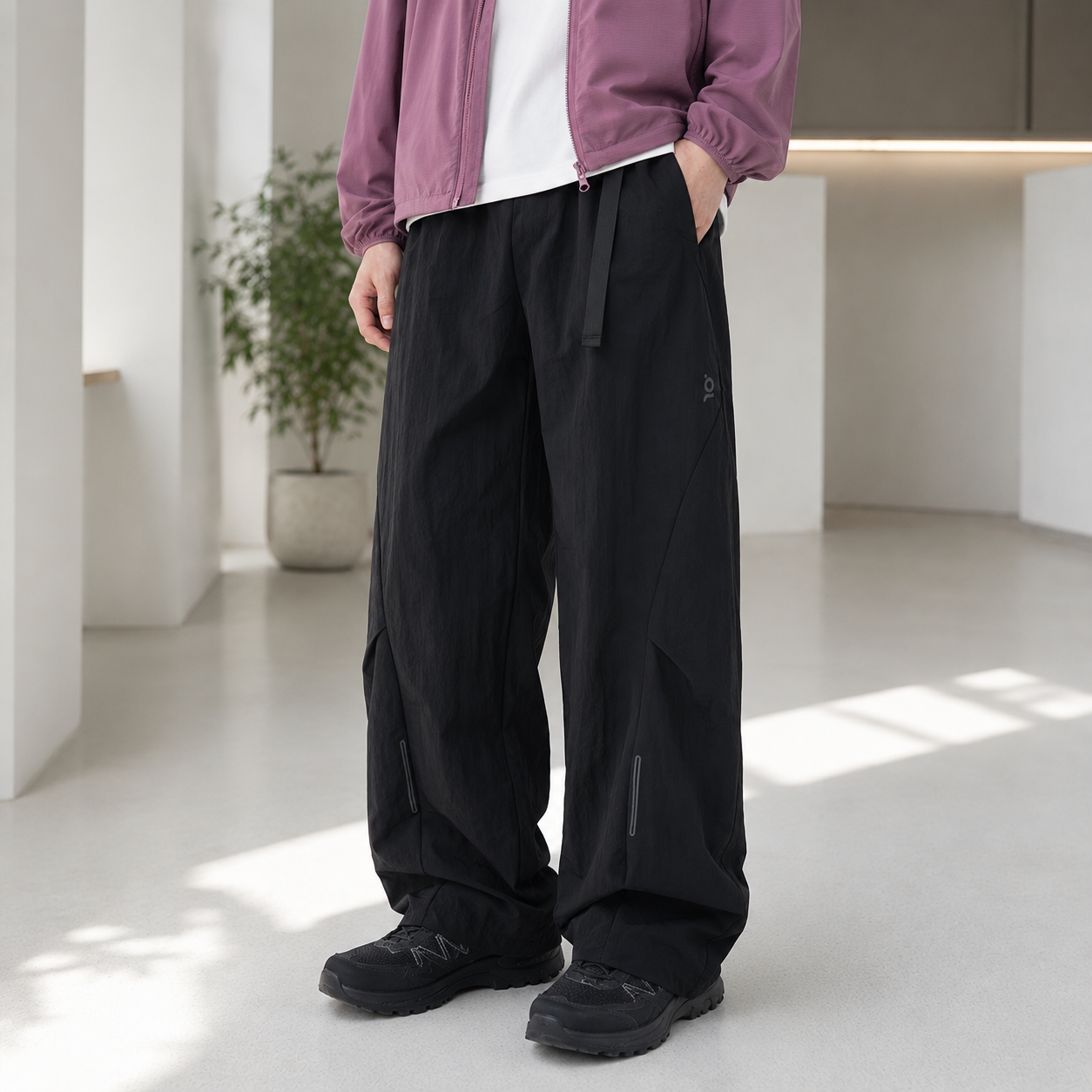 A person wearing loose black wide-leg pants with a drawstring waist, styled with a white t-shirt, a mauve zip-up jacket, and black sneakers in a bright modern indoor setting.
