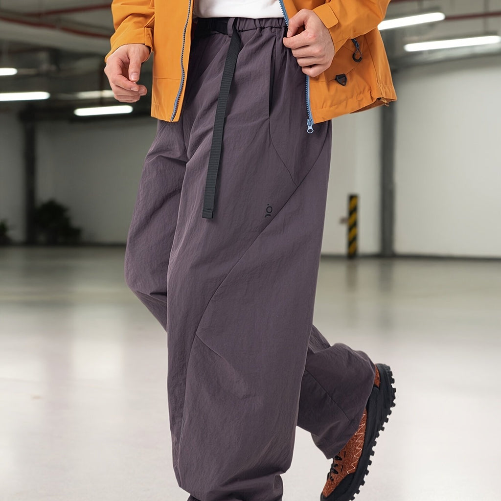 A person wearing loose, oversized dark gray cargo-style pants with a black drawstring, shown from the waist down in an indoor industrial setting. An orange jacket is partially visible.