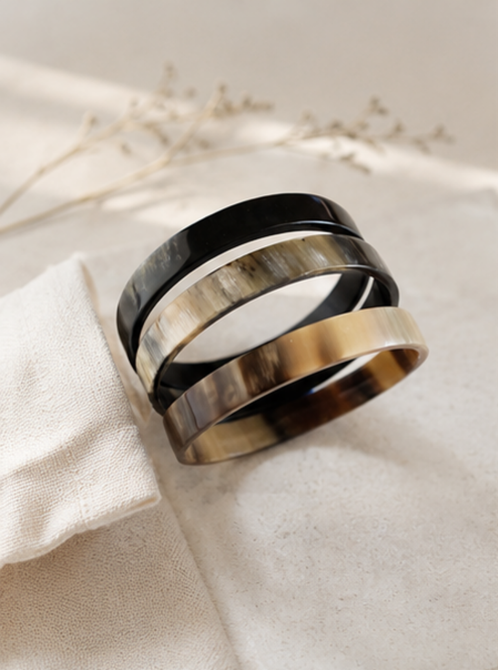 Three wide stacking rings with a glossy tortoiseshell pattern in black, brown, and gold, displayed on a textured cream fabric with dried floral stems in the background.
