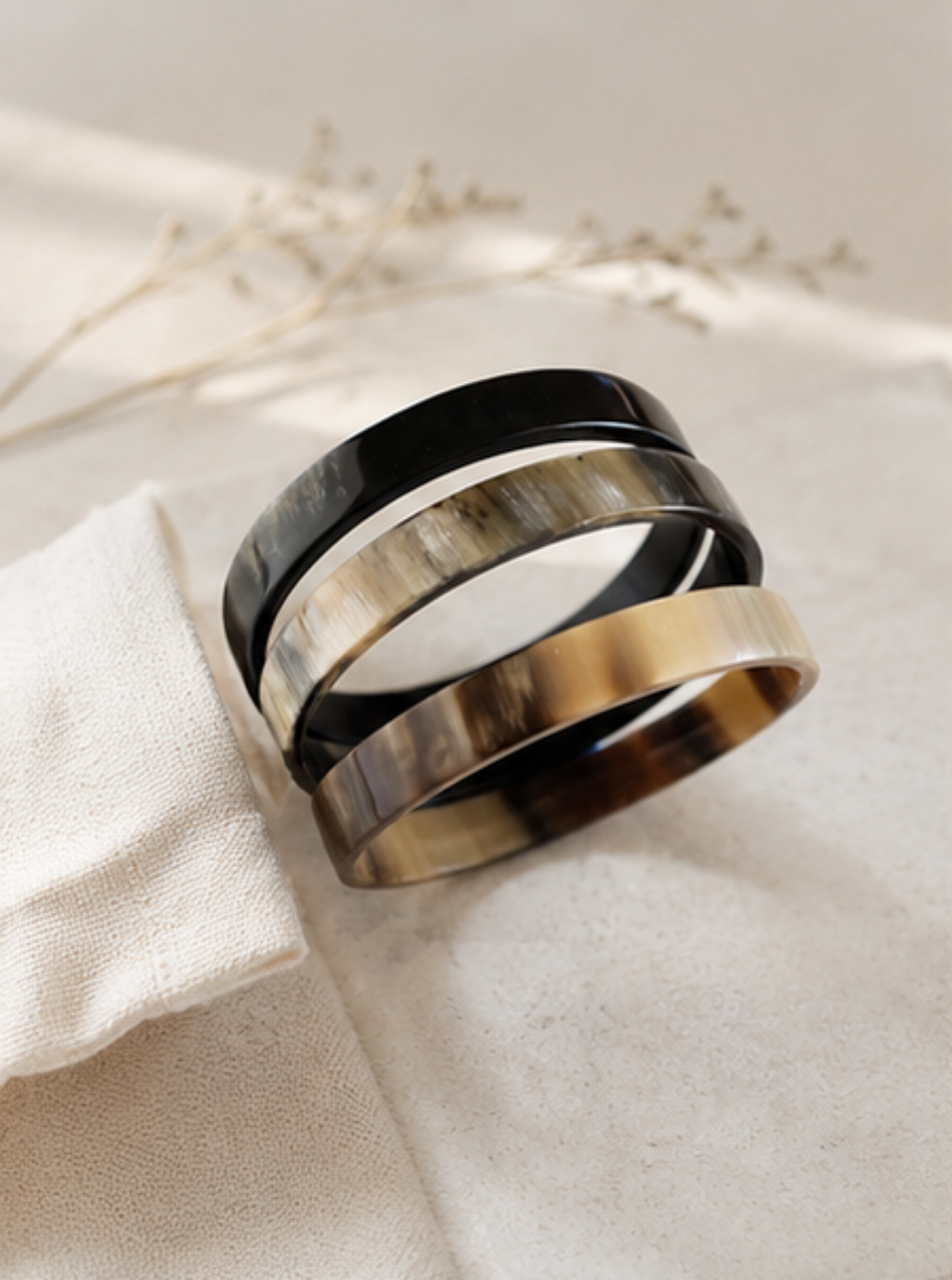 Three wide stacking rings with a glossy tortoiseshell pattern in black, brown, and gold, displayed on a textured cream fabric with dried floral stems in the background.