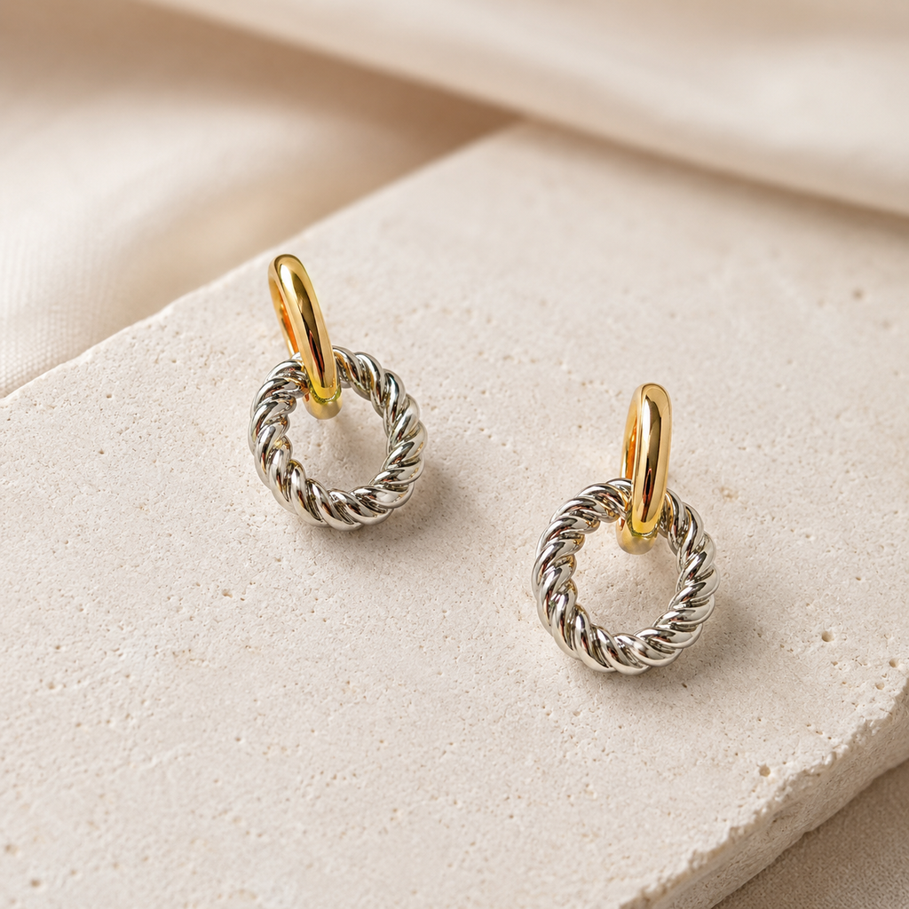 A pair of small hoop earrings with twisted silver-tone wire rings and polished gold-tone posts, displayed on a light beige stone surface with soft cream fabric in the background.