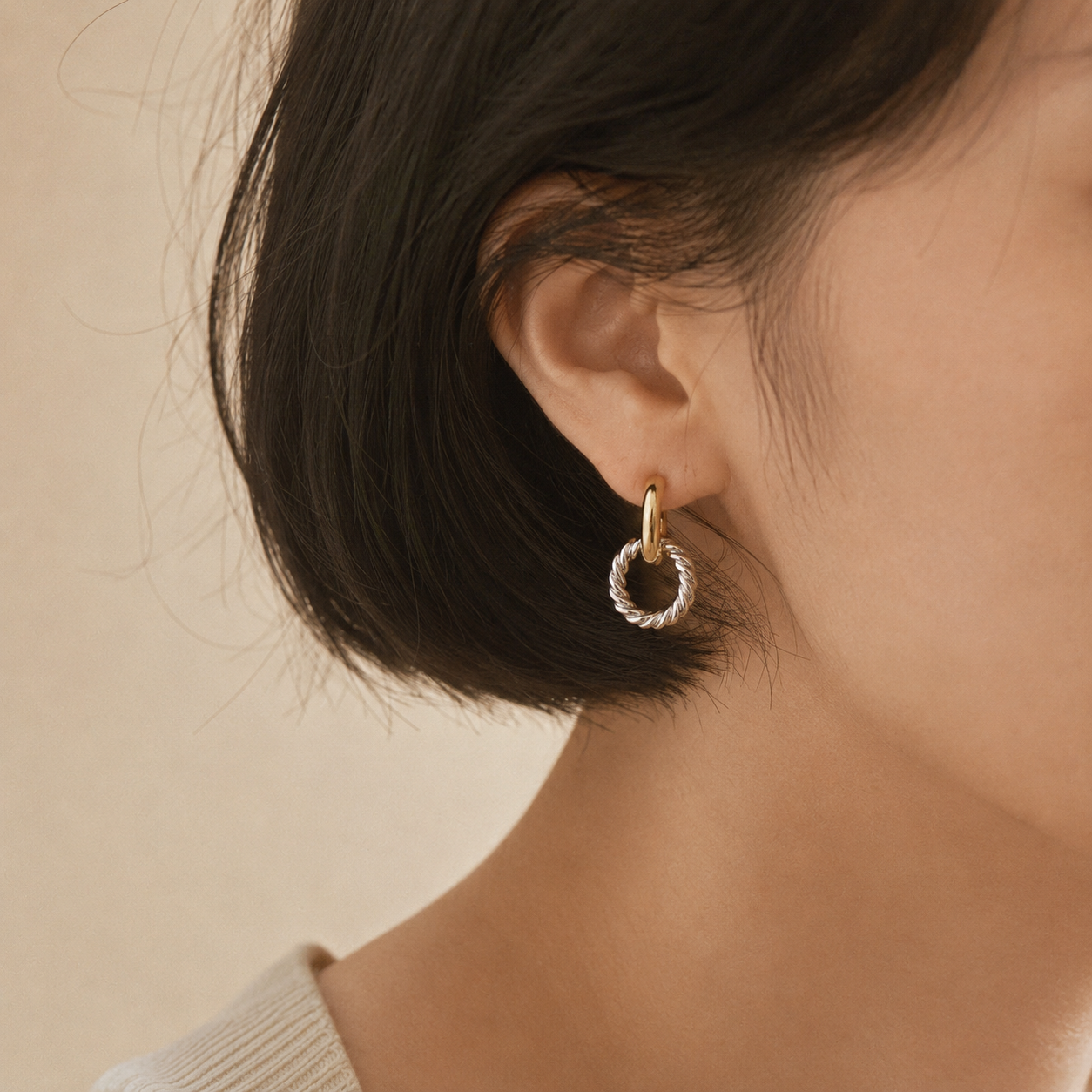 A close-up of a person's ear wearing a small twisted hoop earring in gold and silver tones, against a soft beige background.
