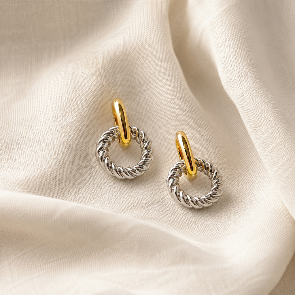 A pair of silver-toned twisted cable hoop earrings with gold-toned ear loops, displayed on a draped cream-colored fabric background.