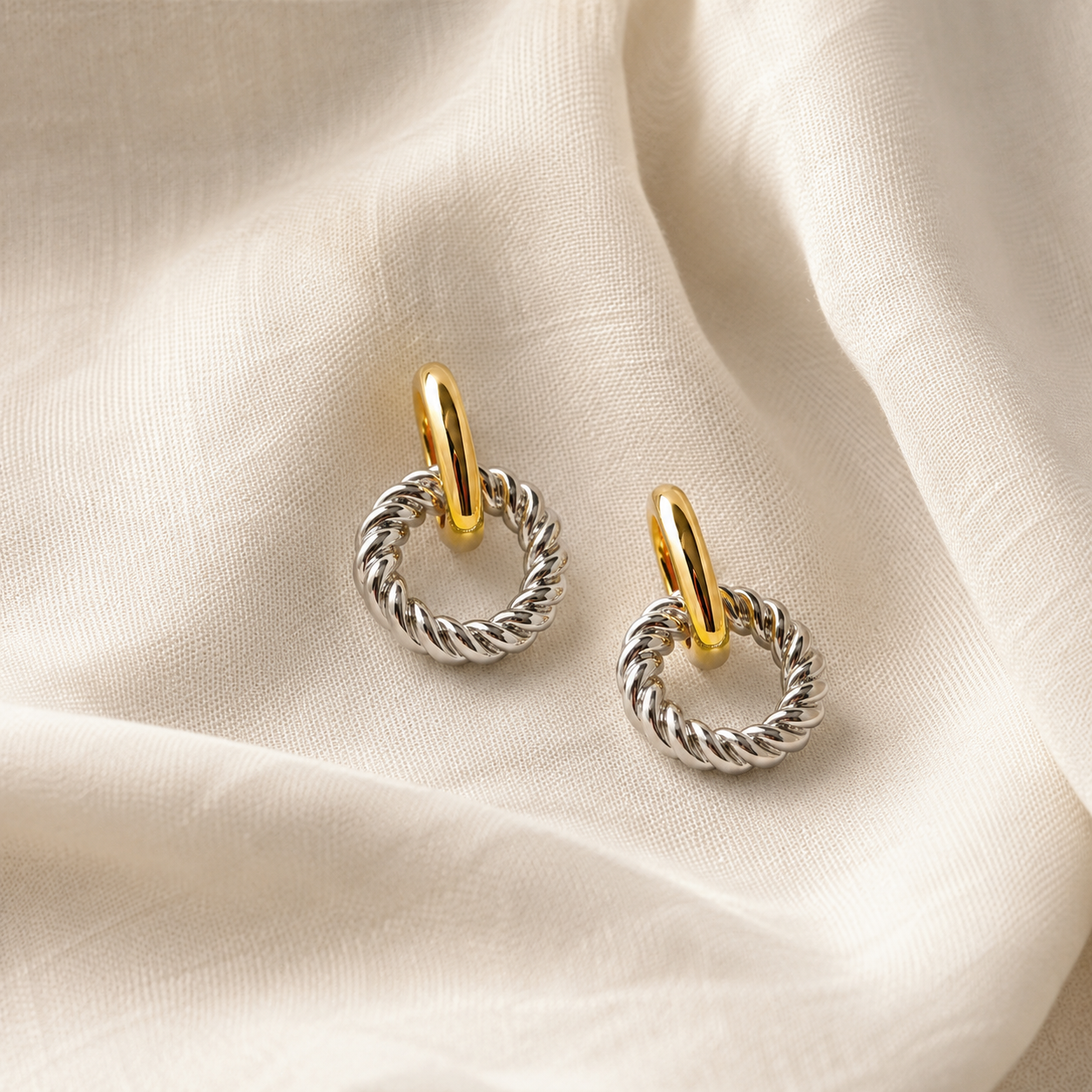 A pair of silver-toned twisted cable hoop earrings with gold-toned ear loops, displayed on a draped cream-colored fabric background.