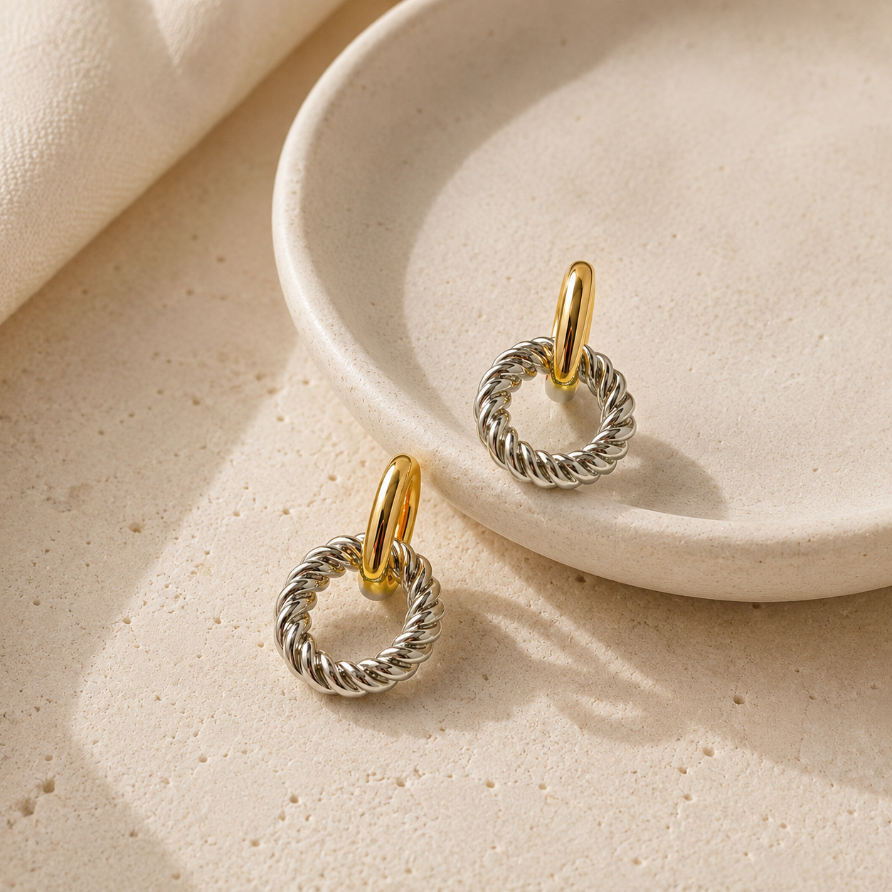 A pair of silver-toned rope-wrapped hoop earrings with gold-toned post tops, displayed on a textured beige surface next to a ceramic dish.