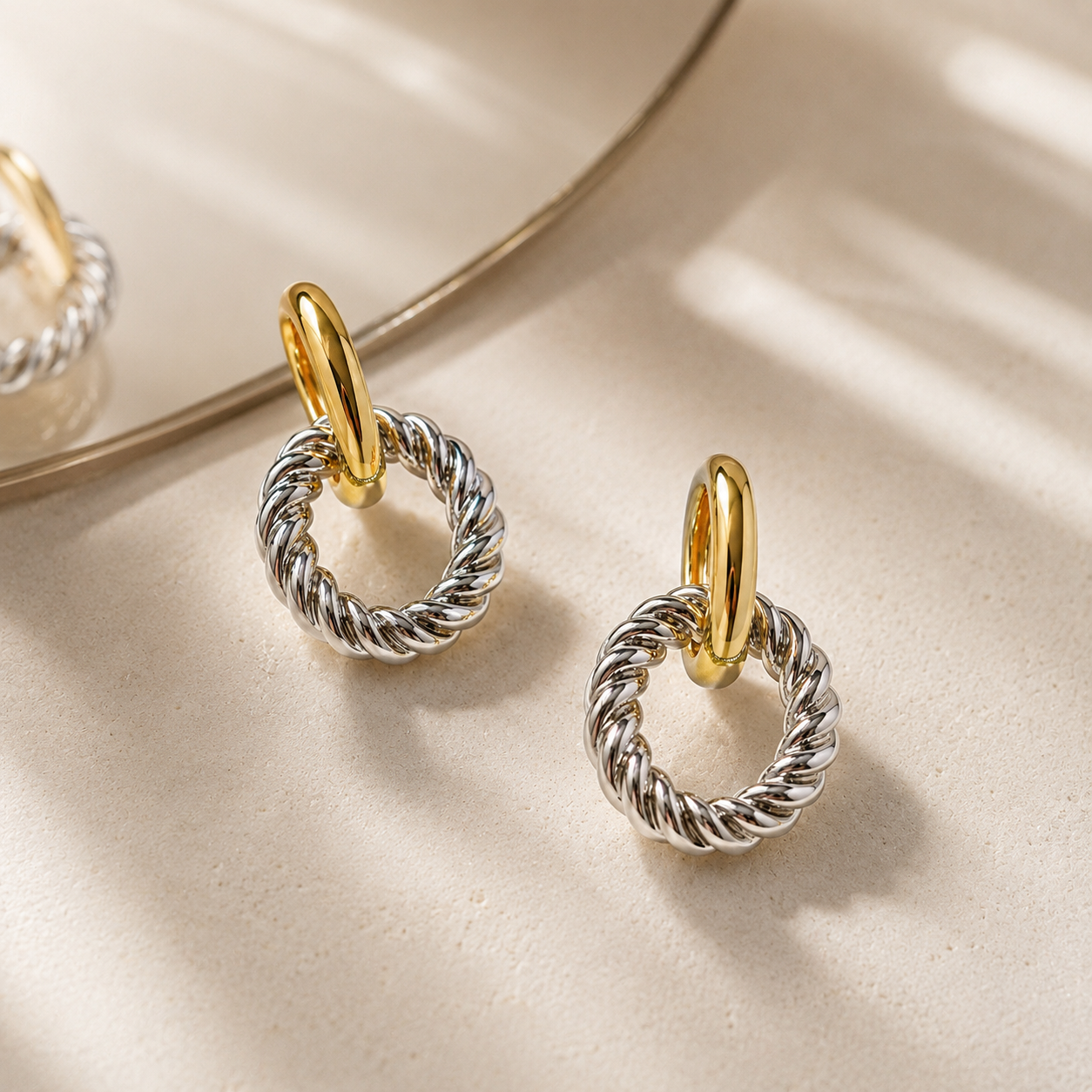 A pair of silver rope-style hoop earrings with gold post connectors, displayed on a textured beige surface with soft shadows and a gold-framed mirror reflection.