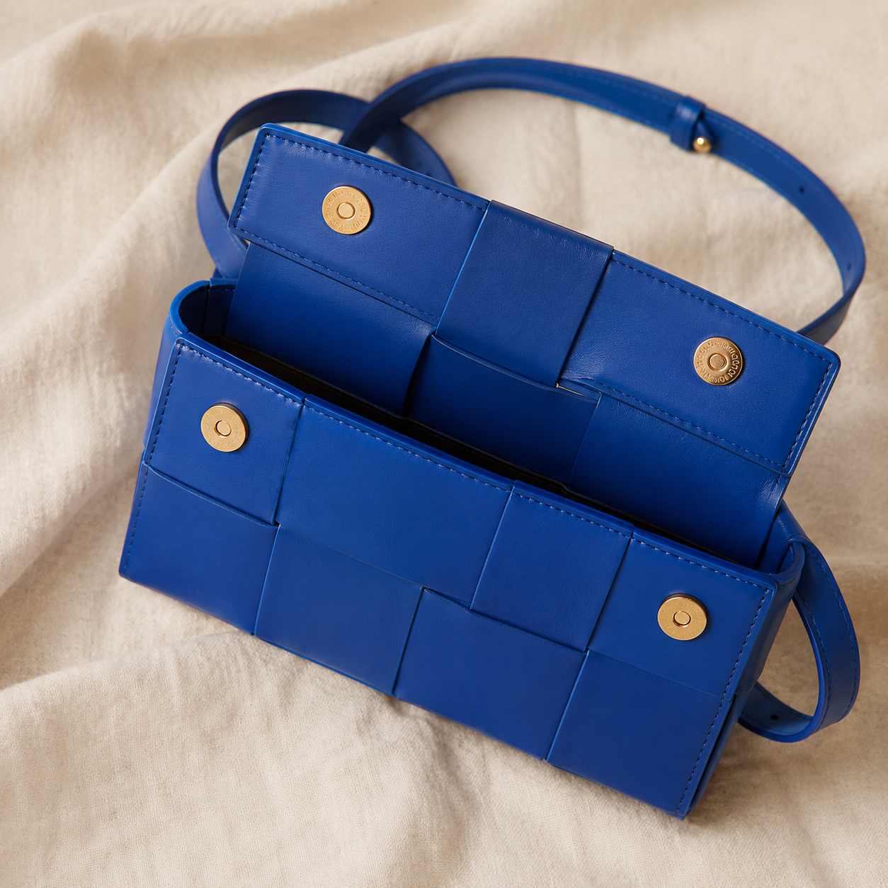 A vibrant royal blue leather shoulder bag with a woven grid pattern and gold-tone snap buttons, displayed on a soft beige fabric background.