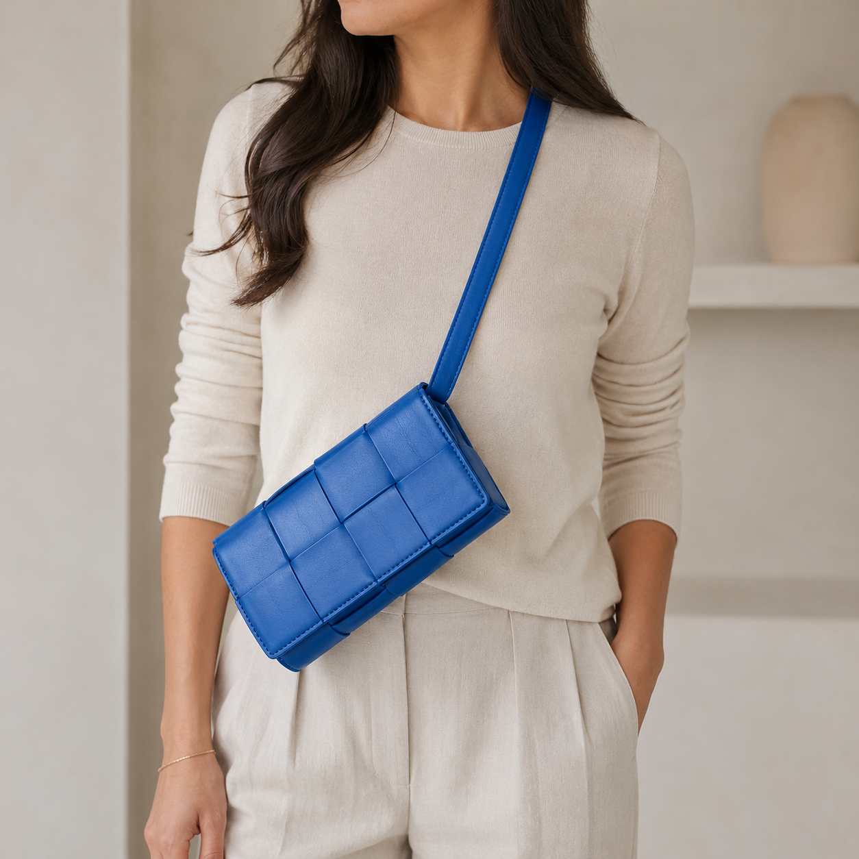 A woman wearing a cream-colored knit sweater and matching pants, carrying a bright blue quilted rectangular crossbody bag with a matching strap.