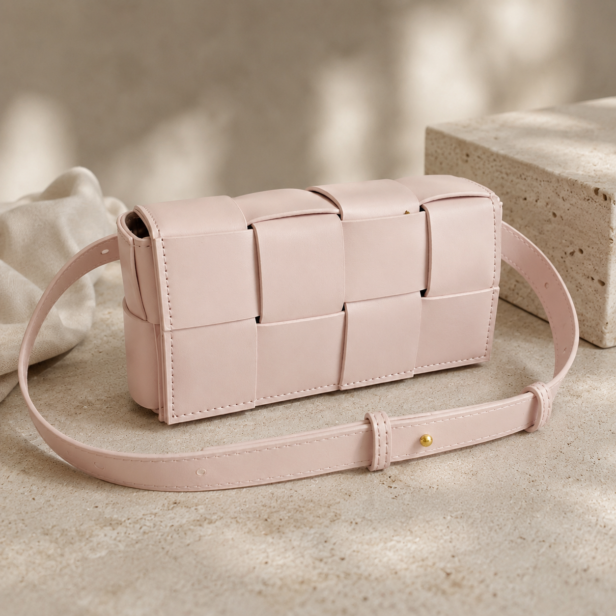 A light pink woven leather shoulder bag with a matching adjustable strap and a small gold-tone buckle, displayed on a textured beige surface with soft neutral background props.
