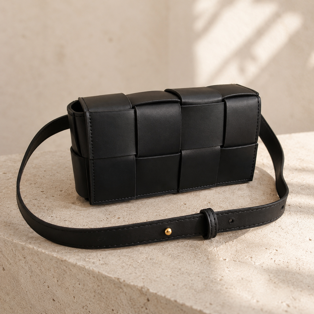 A black woven leather shoulder bag with a long strap, resting on a textured beige stone surface with soft natural shadows.
