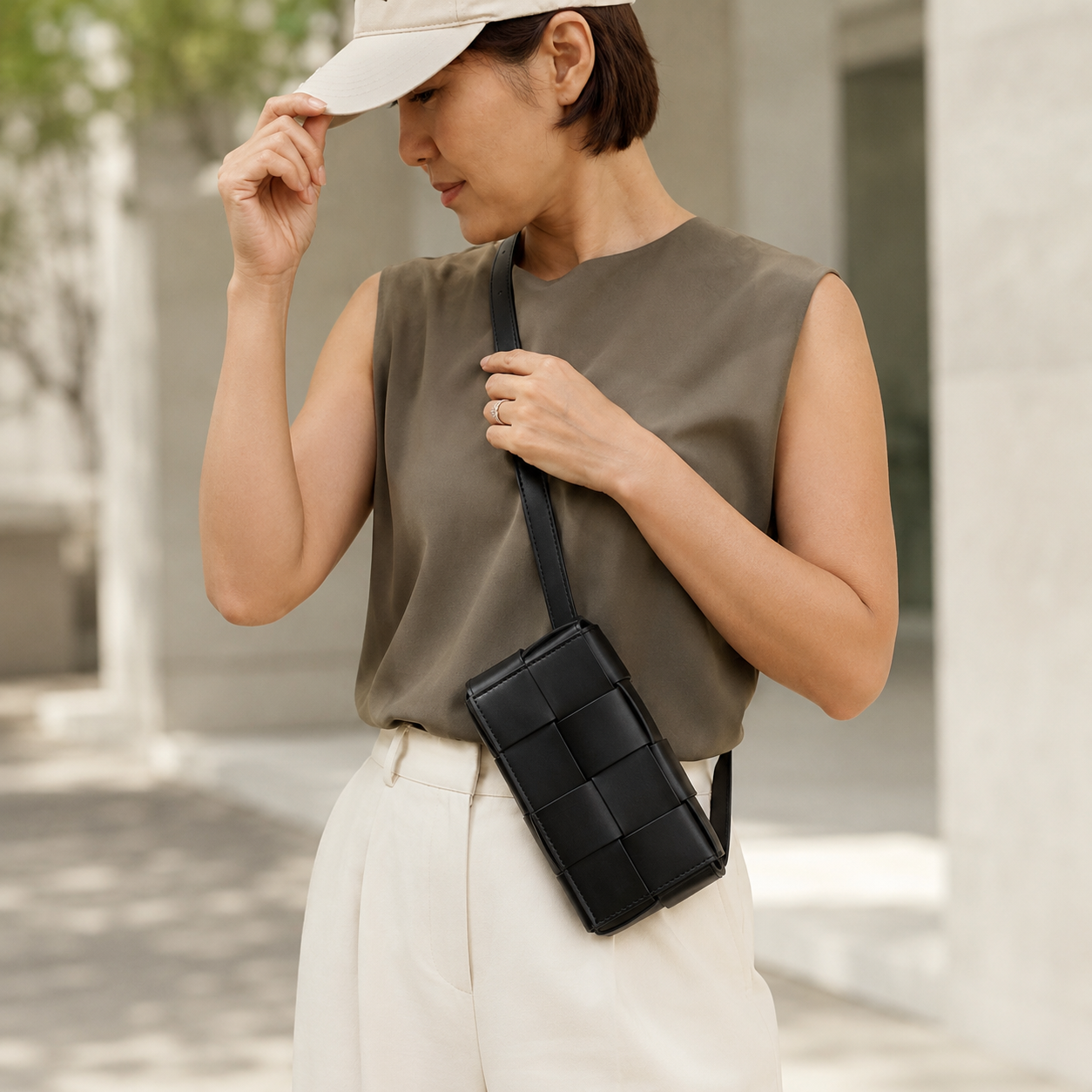 A woman wearing a beige baseball cap, a sleeveless olive green top, and cream-colored trousers carries a black woven crossbody bag with a single strap.